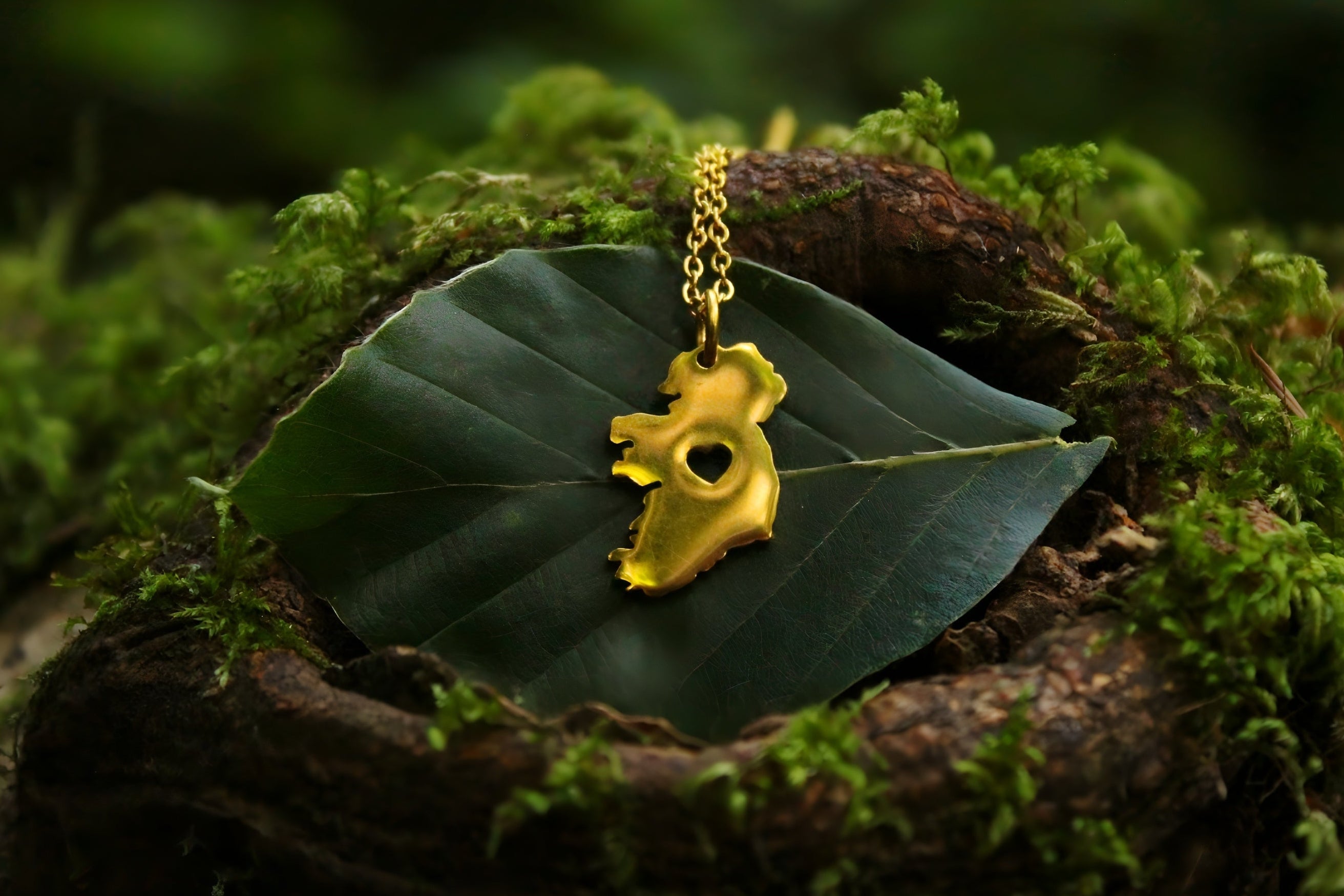 A gold ireland pendant necklace with heart cut out, resting on a green leaf atop a mossy log, with a blurred irish natural forest background.